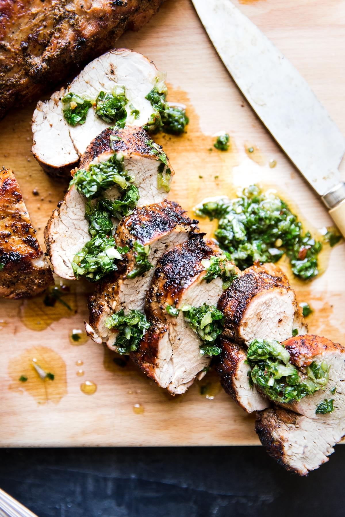 Grilled Pork Tenderloin sliced on a cutting board with a knife with Chimichurri