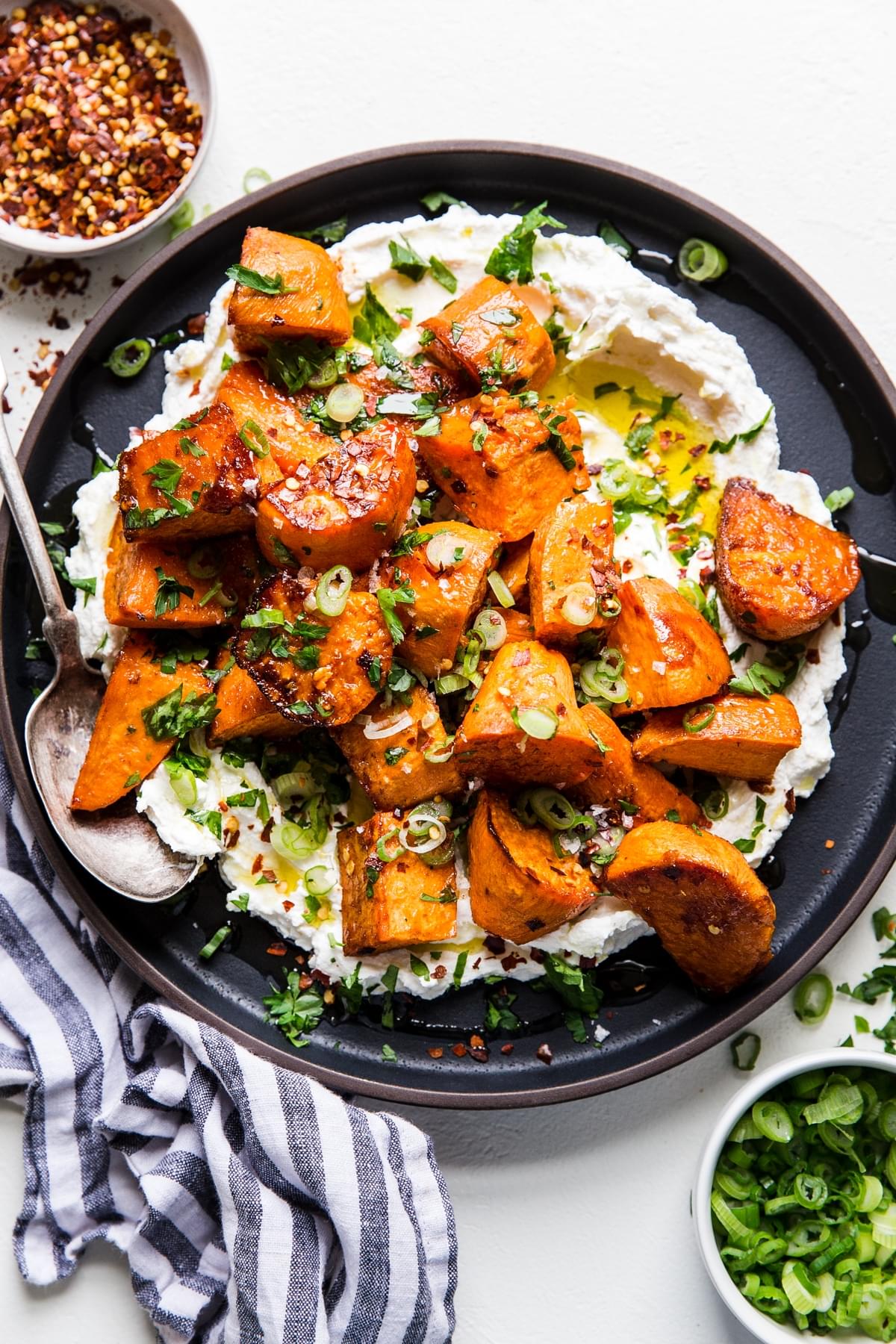 Honey Roasted Sweet Potatoes over labneh with olive oil and green onions on a plate with a linen and serving spoon.
