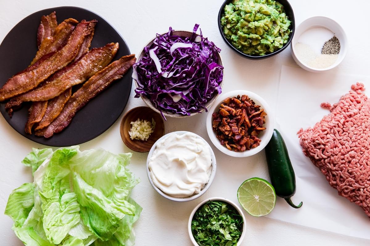 Ingredients laid out bacon, red cabbage, ground beef guacamole, lettuce, jalapeño, garlic.