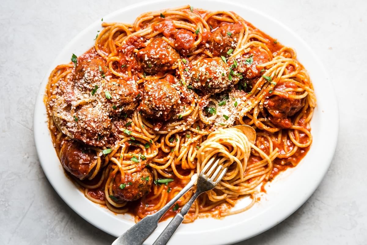 a white platter of cooked spaghetti with homemade tomato sauce and meatballs topped with parmesan cheese and fresh parsley