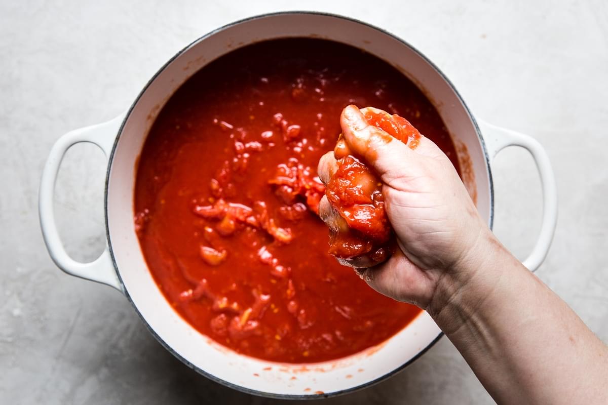 hand crushing whole canned tomatoes into a large pot