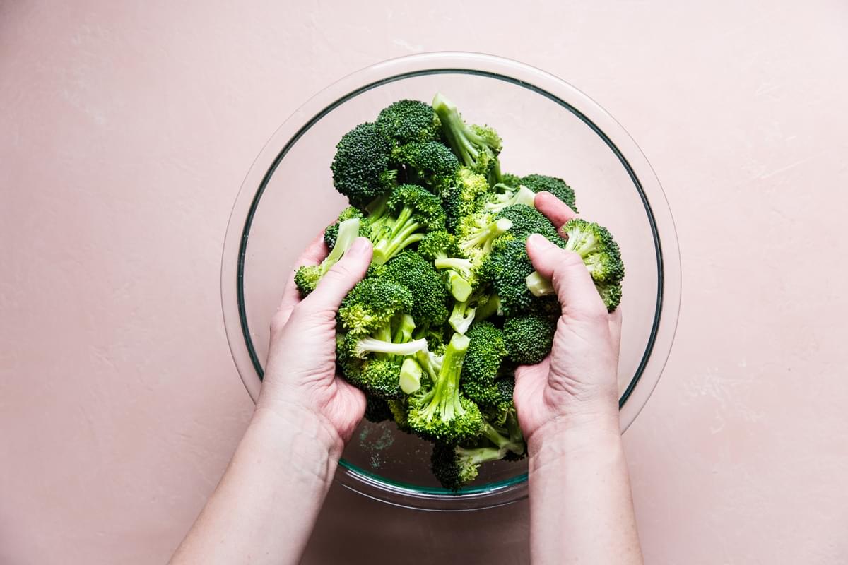 hands putting olive oil on broccoli in a bowl