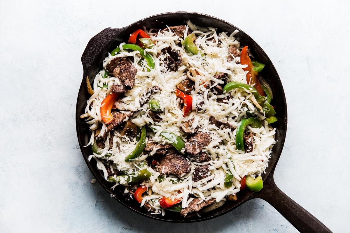 Cheese sprinkled over steak, mushrooms, bell peppers, onions in a pan.
