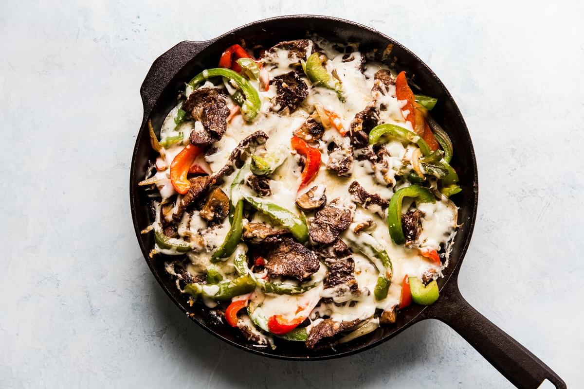 Cheese melted over steak, mushrooms, bell peppers, onions in a pan.