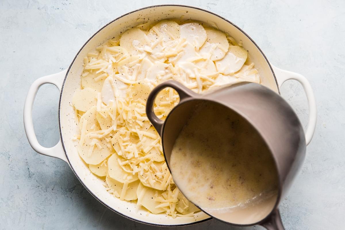 butter and cream sauce being poured over layers of sliced potatoes and Gruyère cheese in a casserole dish