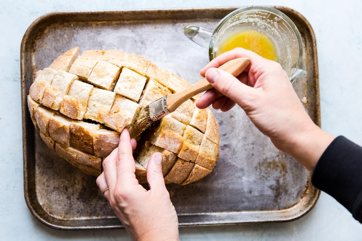 hands brushing garlic butter into sliced crusty bread