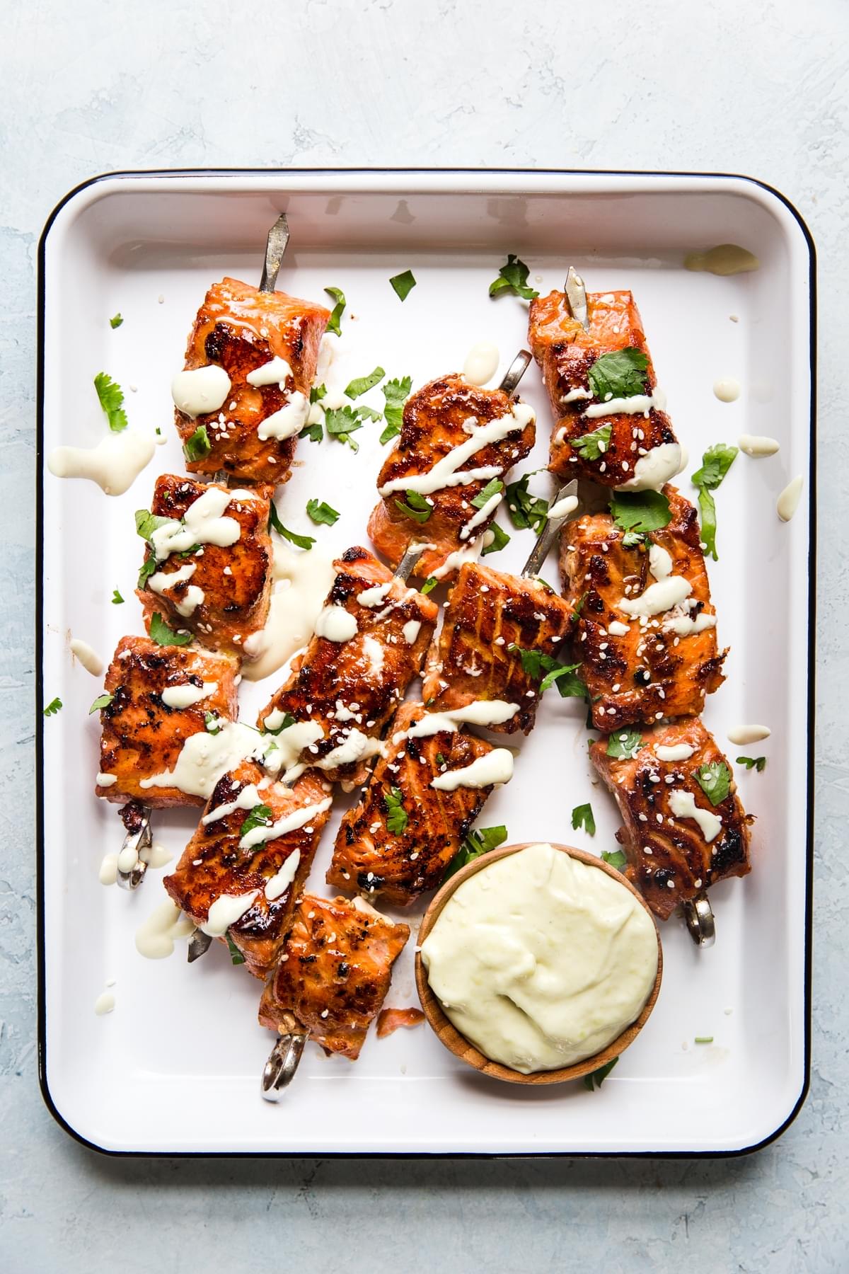 salmon bite skewers on a white metal tray drizzled with wasabi aioli and topped with fresh cilantro and sesame seeds