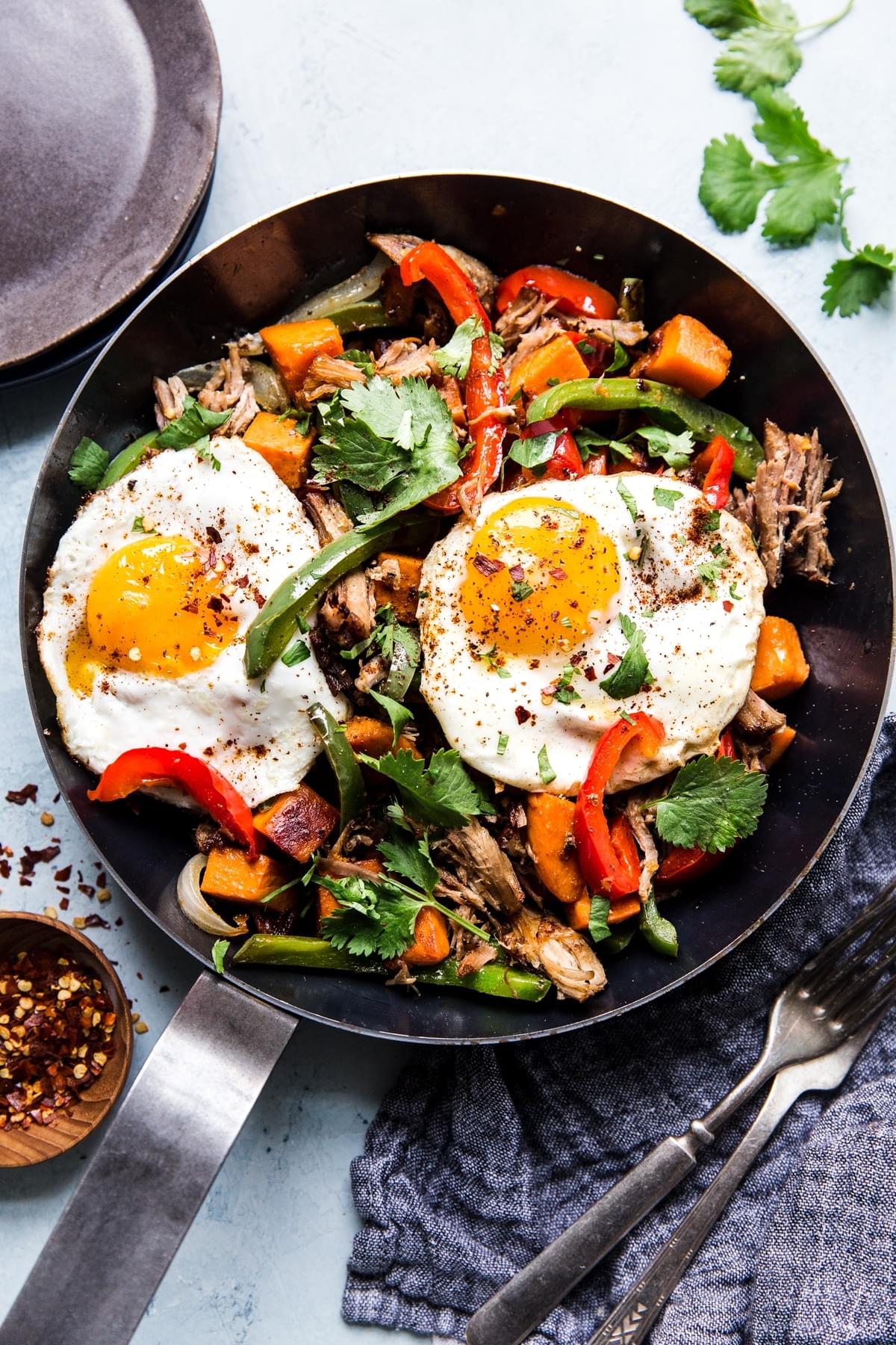 Sweet Potato Carnitas Hash in a pan with red and green bell peppers 2 fried eggs and cilantro