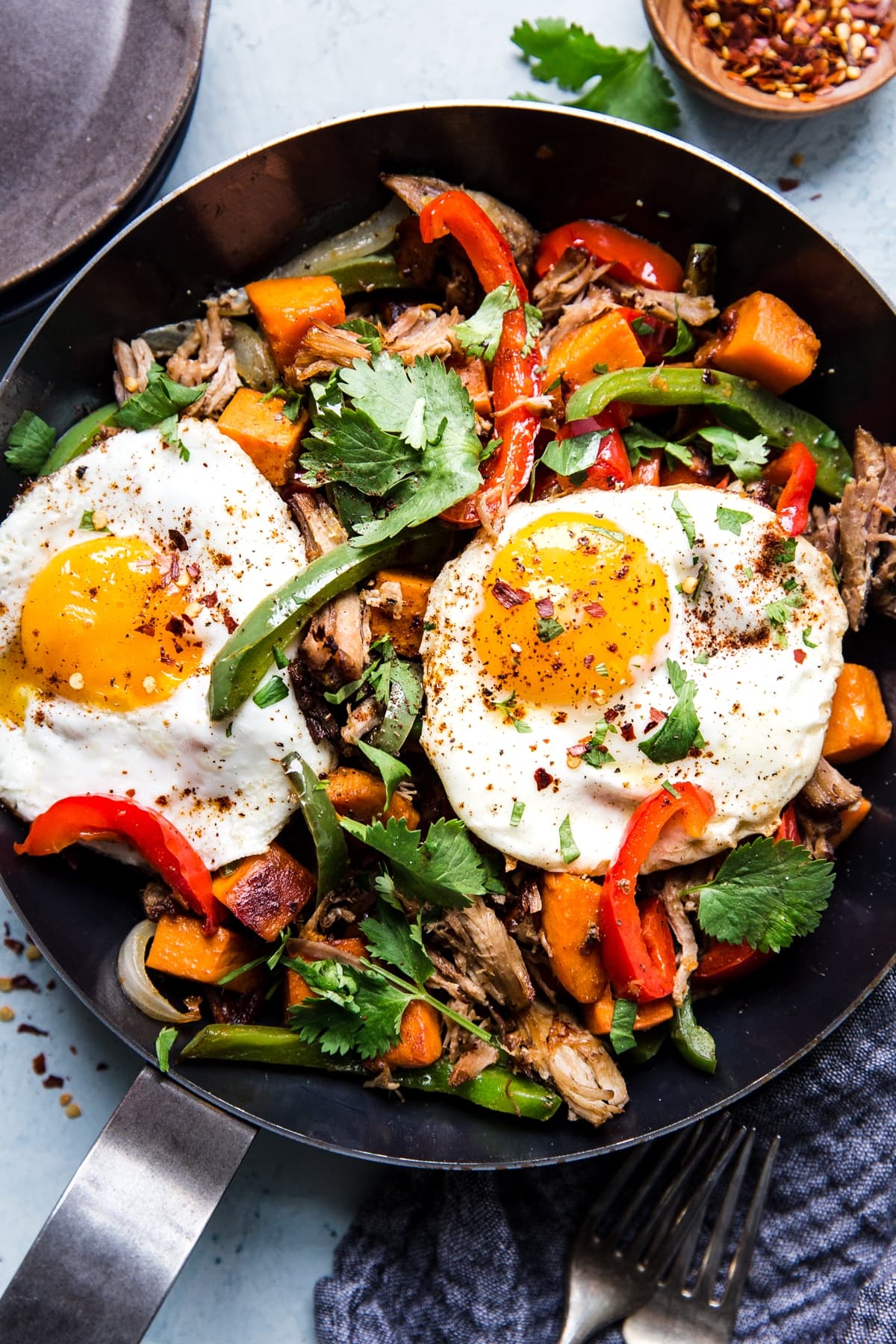 Sweet Potato Carnitas Hash in a pan with red and green bell peppers 2 fried eggs and cilantro