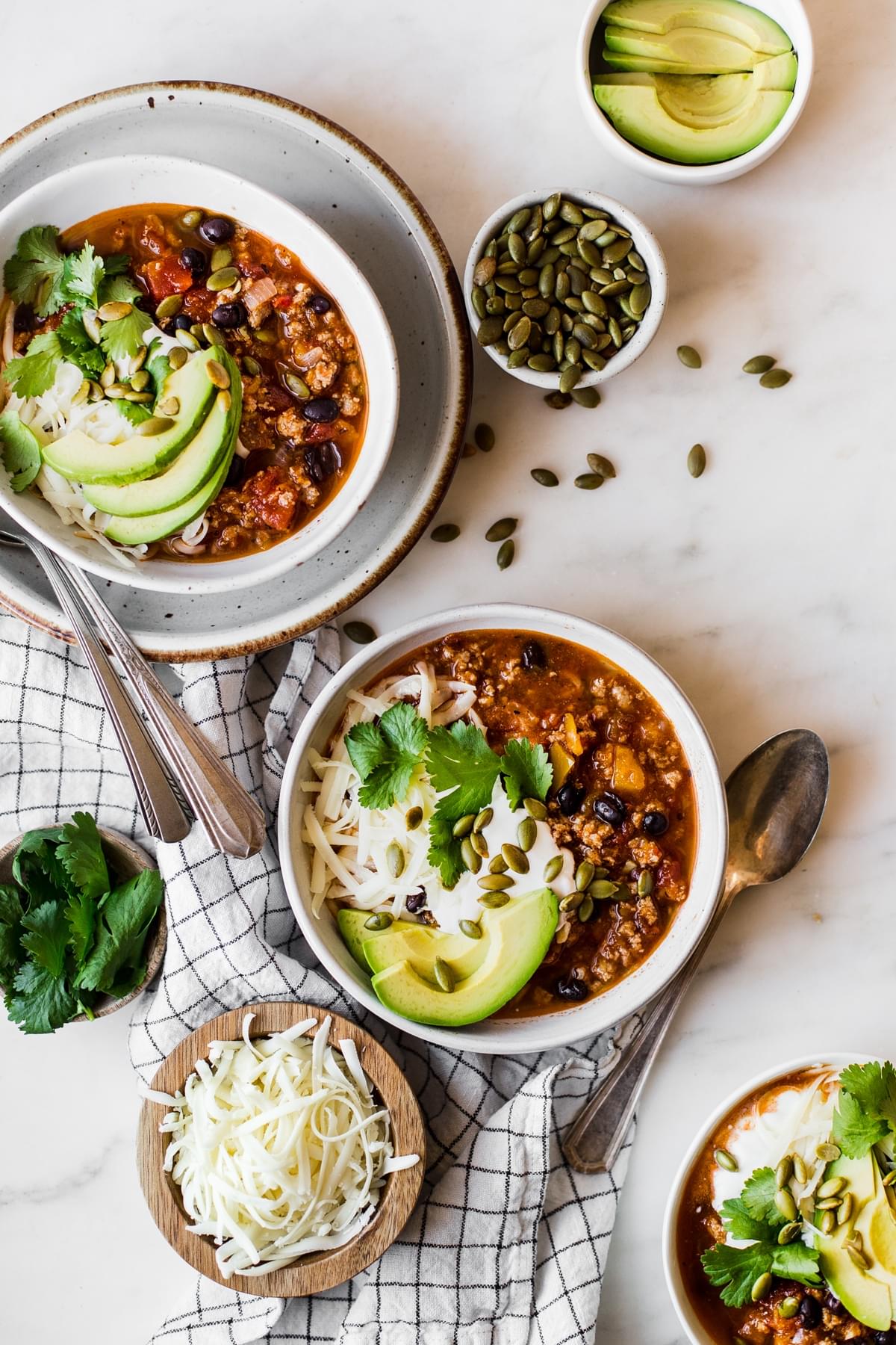 three bowls of turkey pumpkin chili surrounded by small bowls of fresh cilantro, avocado, grated cheese and pumpkin seeds