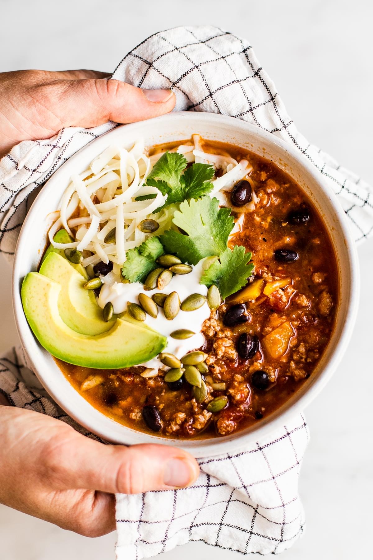 hands holding a bowl of turkey pumpkin chili topped with avocado, grated cheese, fresh cilantro and pumpkin seeds