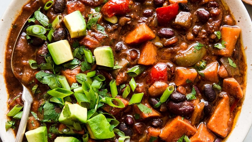 a bowl of homemade vegetarian chili made with bell pepper, sweet potato, onion and beans topped with avocado and green onions