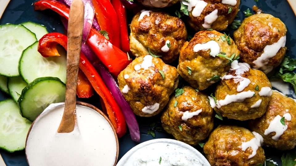 chicken shawarma meatballs on a blue plate shown with roasted red peppers, onions, cucumbers, tahini and tzatziki sauce.