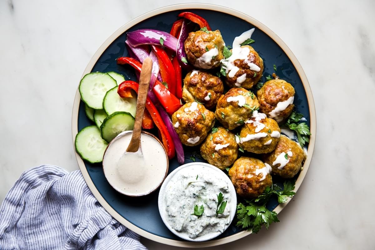 chicken shawarma meatballs on a blue plate with small bowls of tzatziki, tahini and cucumbers.
