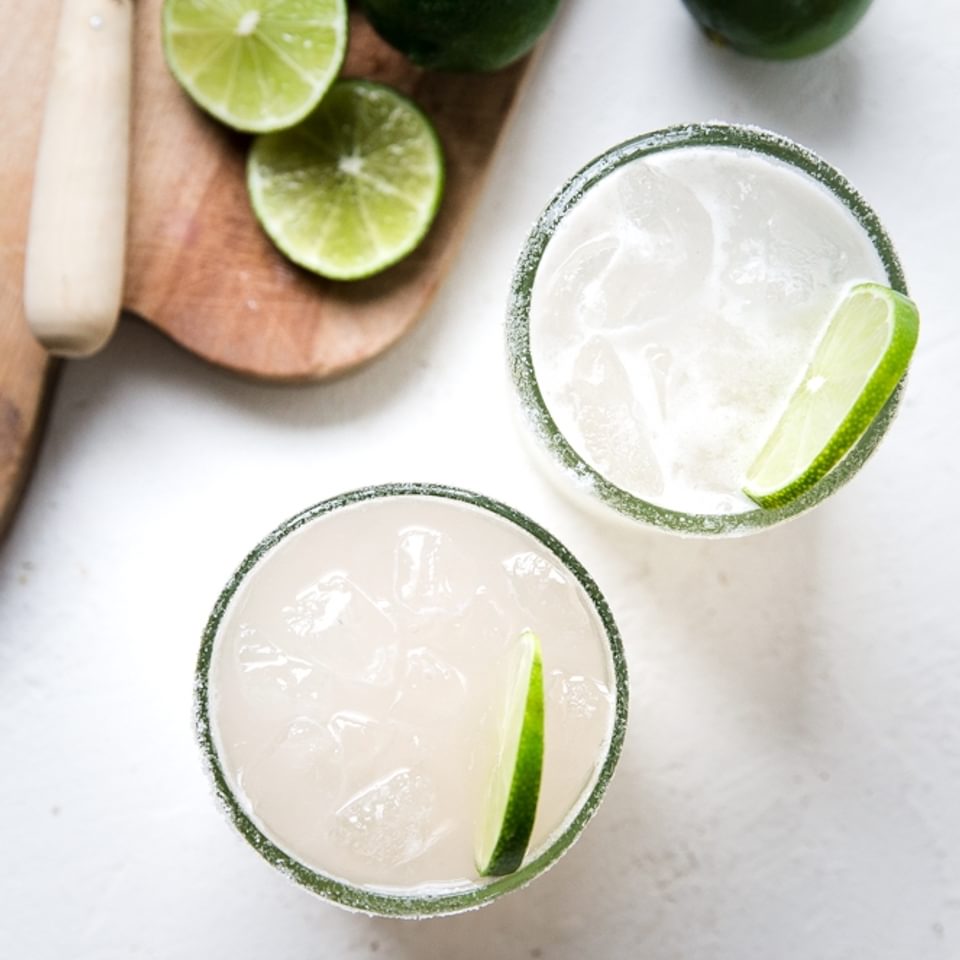 two classic margaritas in glass cups next to a wooden cutting board with sliced limes