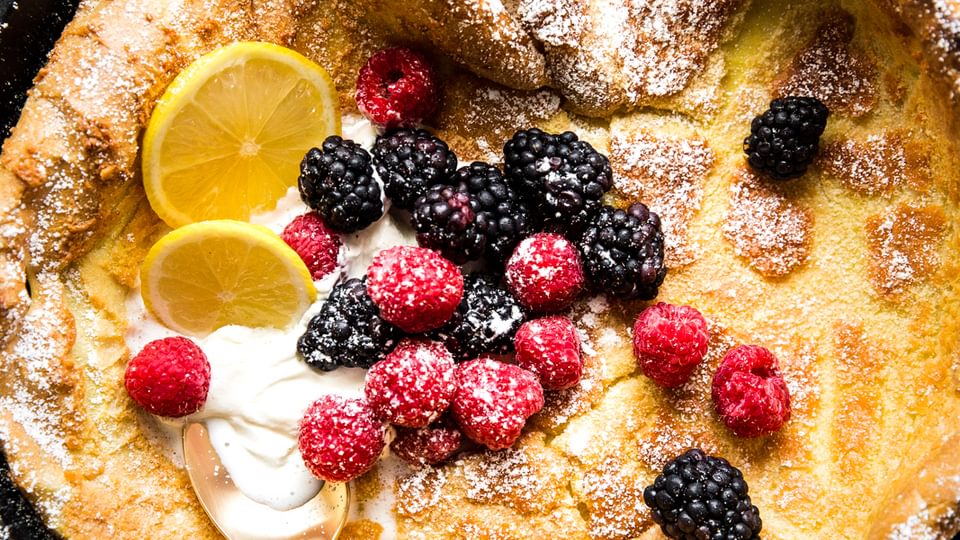 Dutch baby pancake in a cast iron skillet with fresh berries, butter, lemon and maple syrup