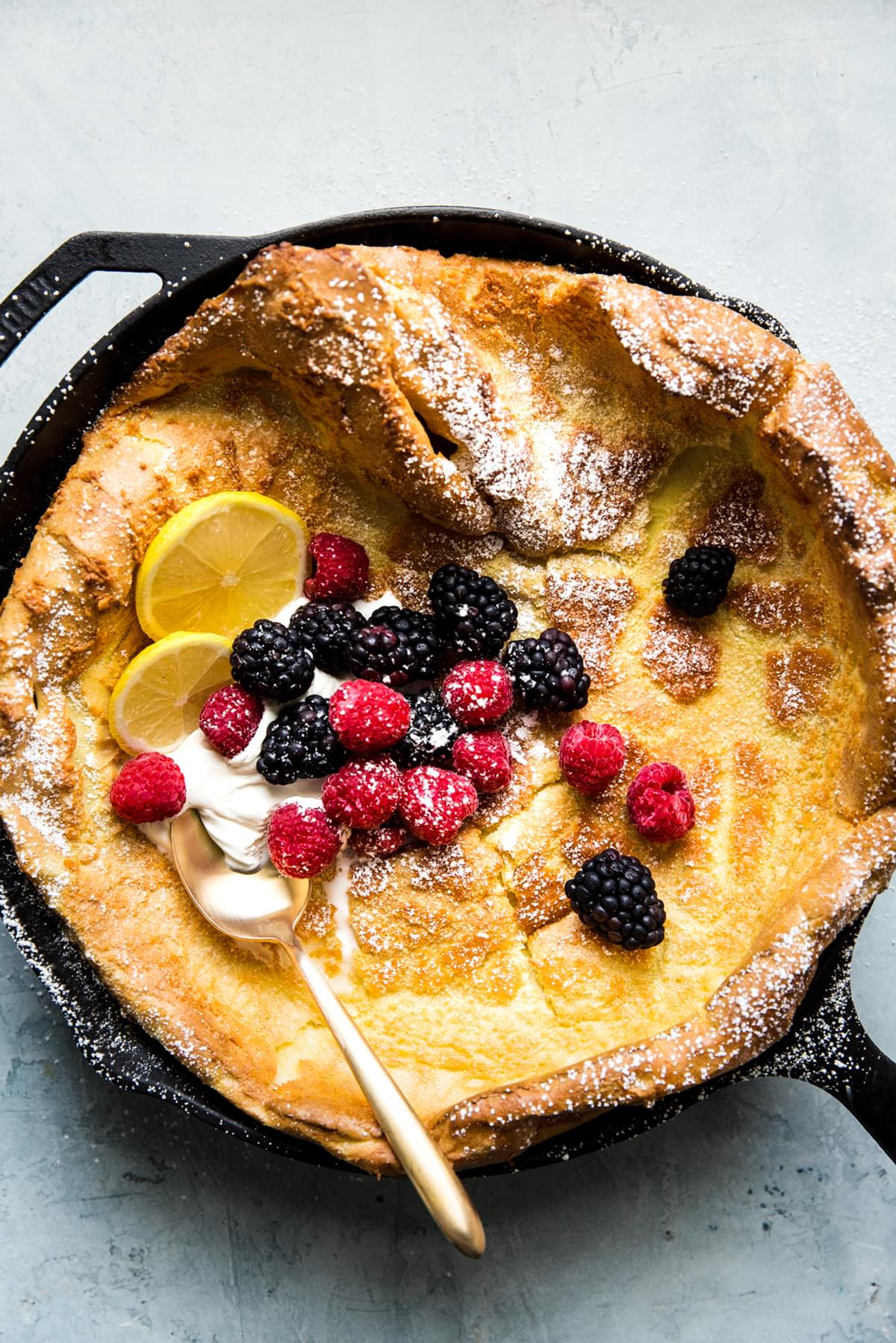 Homemade Dutch baby pancake in a cast iron skillet topped with whipped cream, powdered sugar, berries and lemon