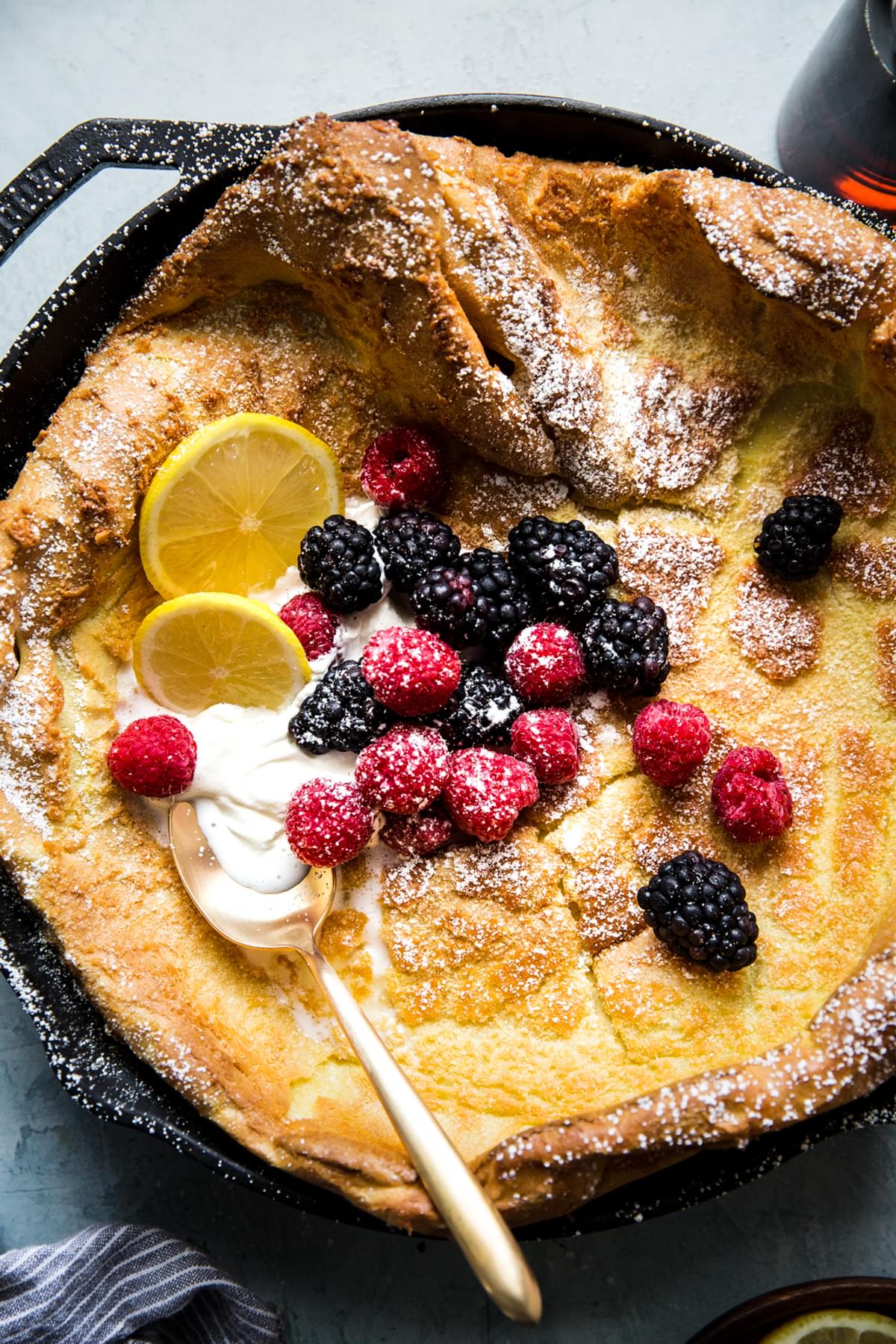 Homemade Dutch baby pancake  in a cast iron skillet topped with whipped cream, powdered sugar, berries and lemon