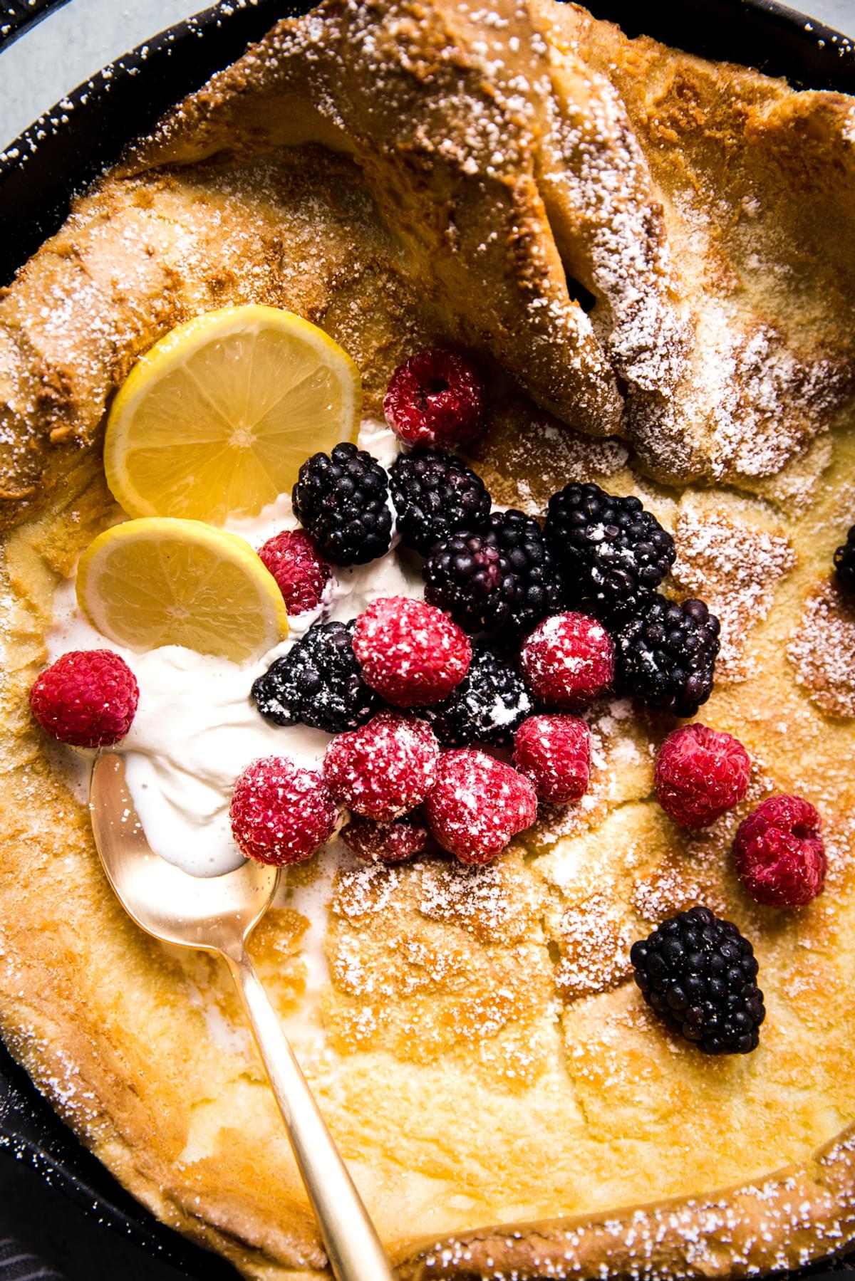 Homemade Dutch baby pancake in a cast iron skillet topped with whipped cream, powdered sugar, berries and lemon
