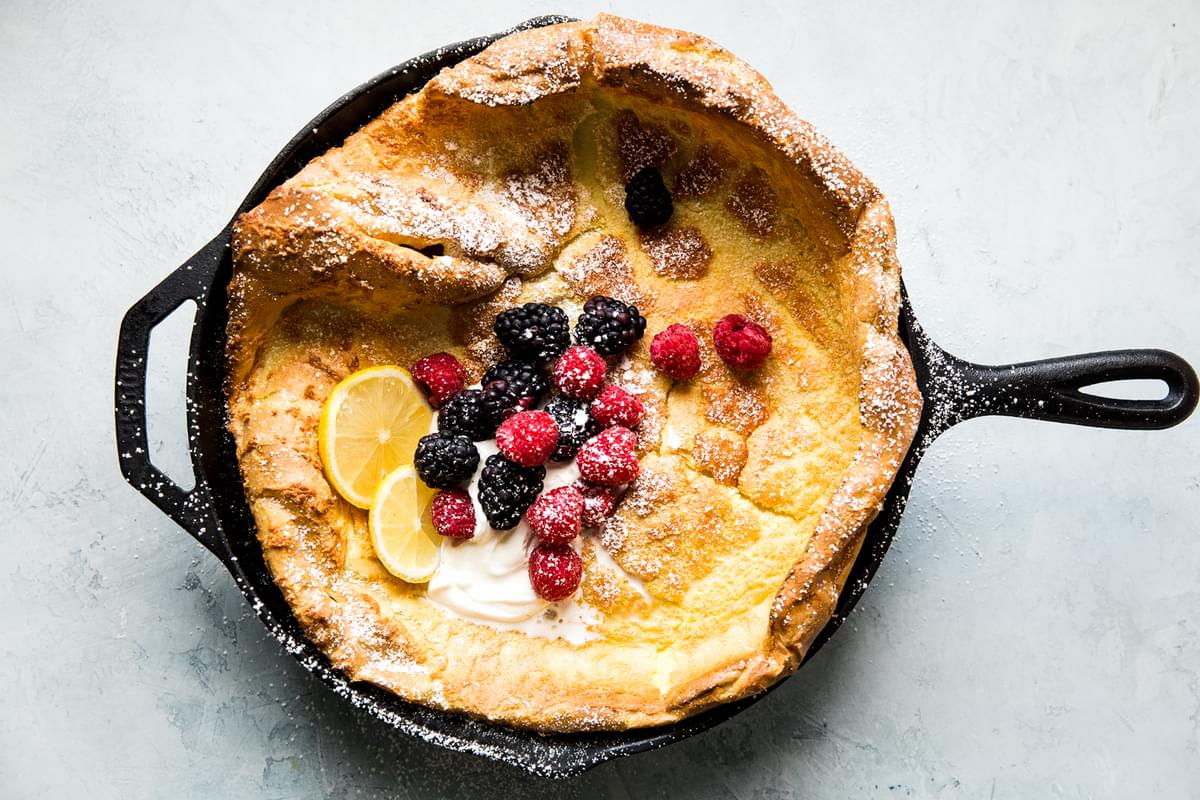 Homemade Dutch baby pancake  in a cast iron skillet topped with whipped cream, powdered sugar, berries and lemon