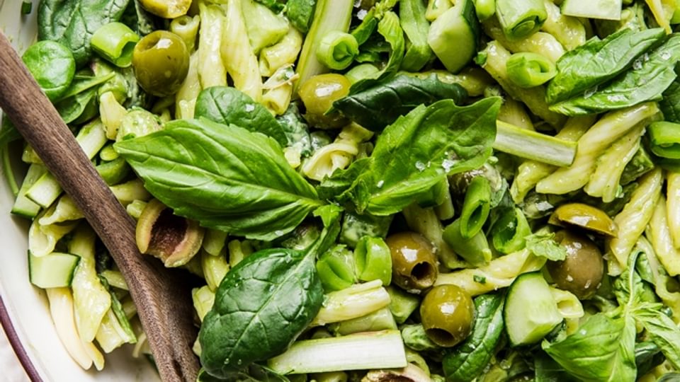 Green goddess pasta salad with basil, green olives, asparagus, snap peas and cucumbers in a a bowl with a wooden spoon