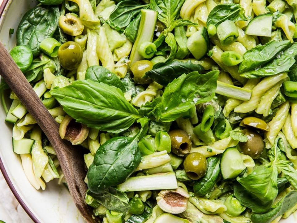 Green goddess pasta salad with basil, green olives, asparagus, snap peas and cucumbers in a a bowl with a wooden spoon