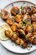 A plate of grilled scallops on metal skewers with lemon slices and parsley