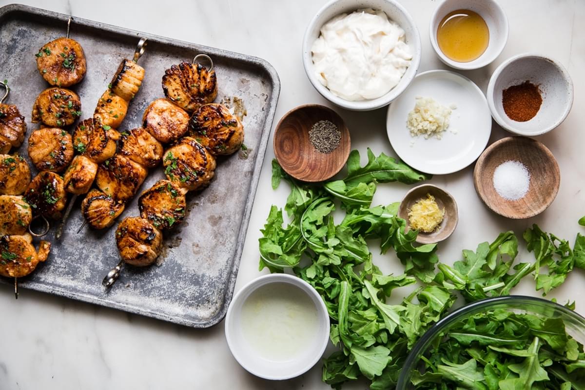 grilled scallops on a baking sheet next to arugula and the ingredients to make lemon aioli