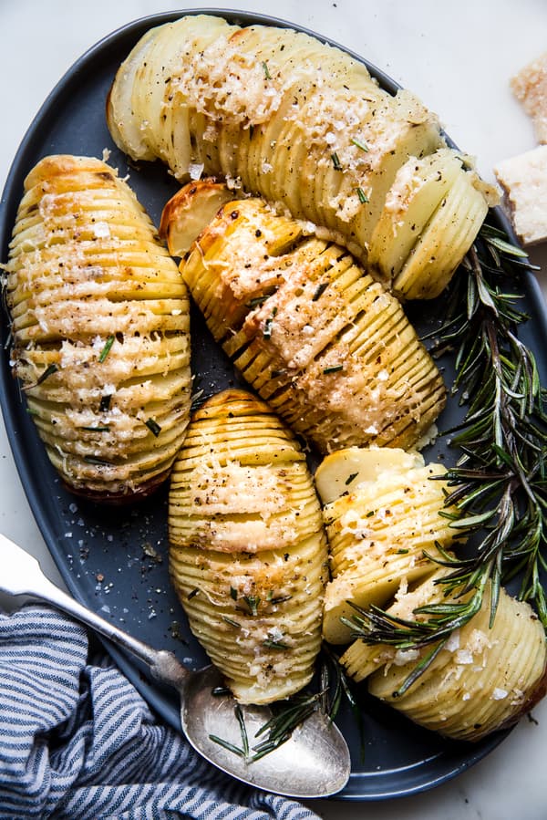 5 hasselback potatoes shown on a large platter with parmesan and fresh rosemary