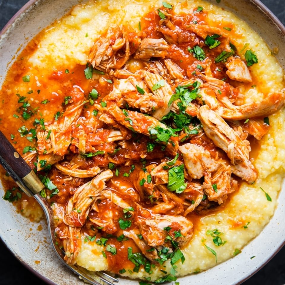 Shredded chicken thighs covered with marinara over polenta in a bowl with fresh basil