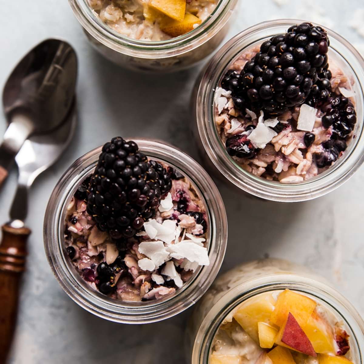 Peaches and Cream overnight oats next to blackberry overnight oats in glass jars next to two spoons.