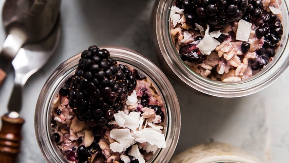Peaches and Cream overnight oats next to blackberry overnight oats in glass jars next to two spoons.