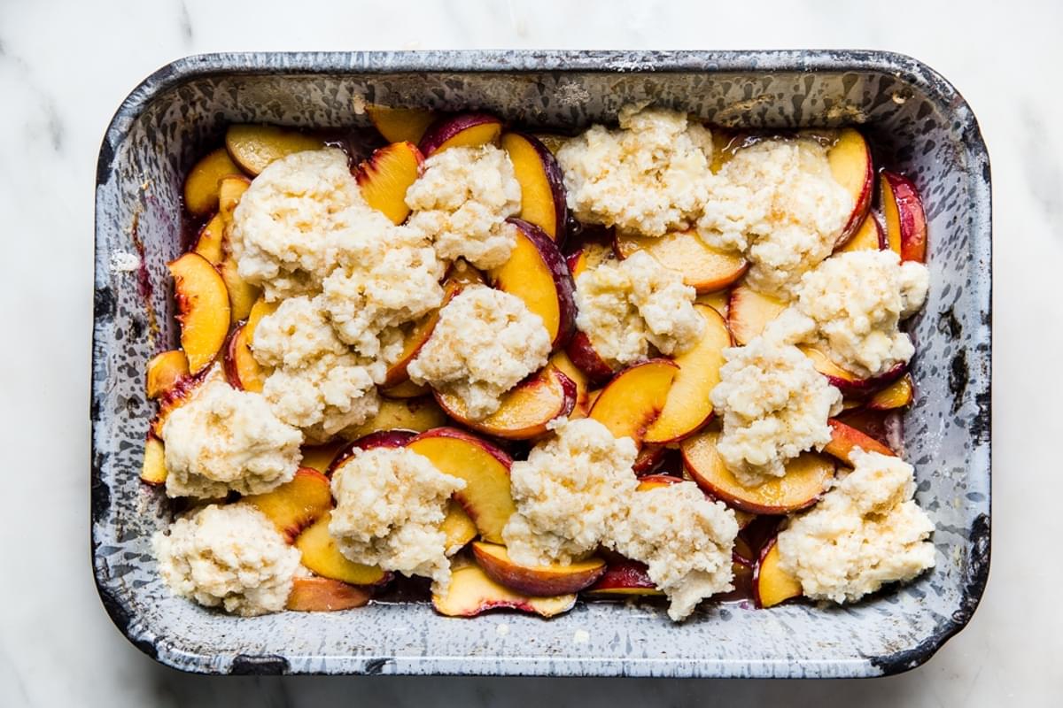 cooked sliced yellow peaches in a baking tin topped with raw cobbler dough getting ready to bake in the oven.