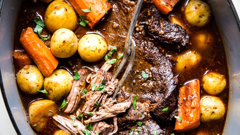 Pot roast shown in a large green dutch oven topped with fresh parsley and served with baby potatoes and carrots