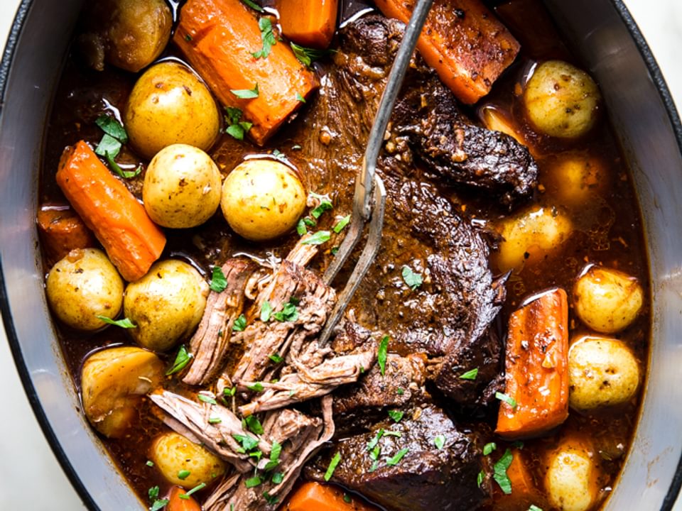 Pot roast shown in a large green dutch oven topped with fresh parsley and served with baby potatoes and carrots