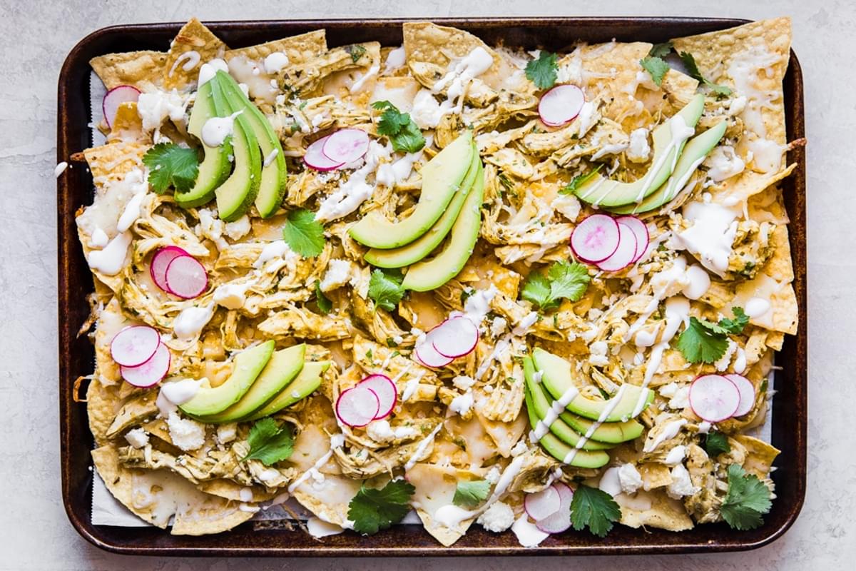 salsa verde nachos topped with sour cream, cilantro, avocado and radishes shown on a baking sheet