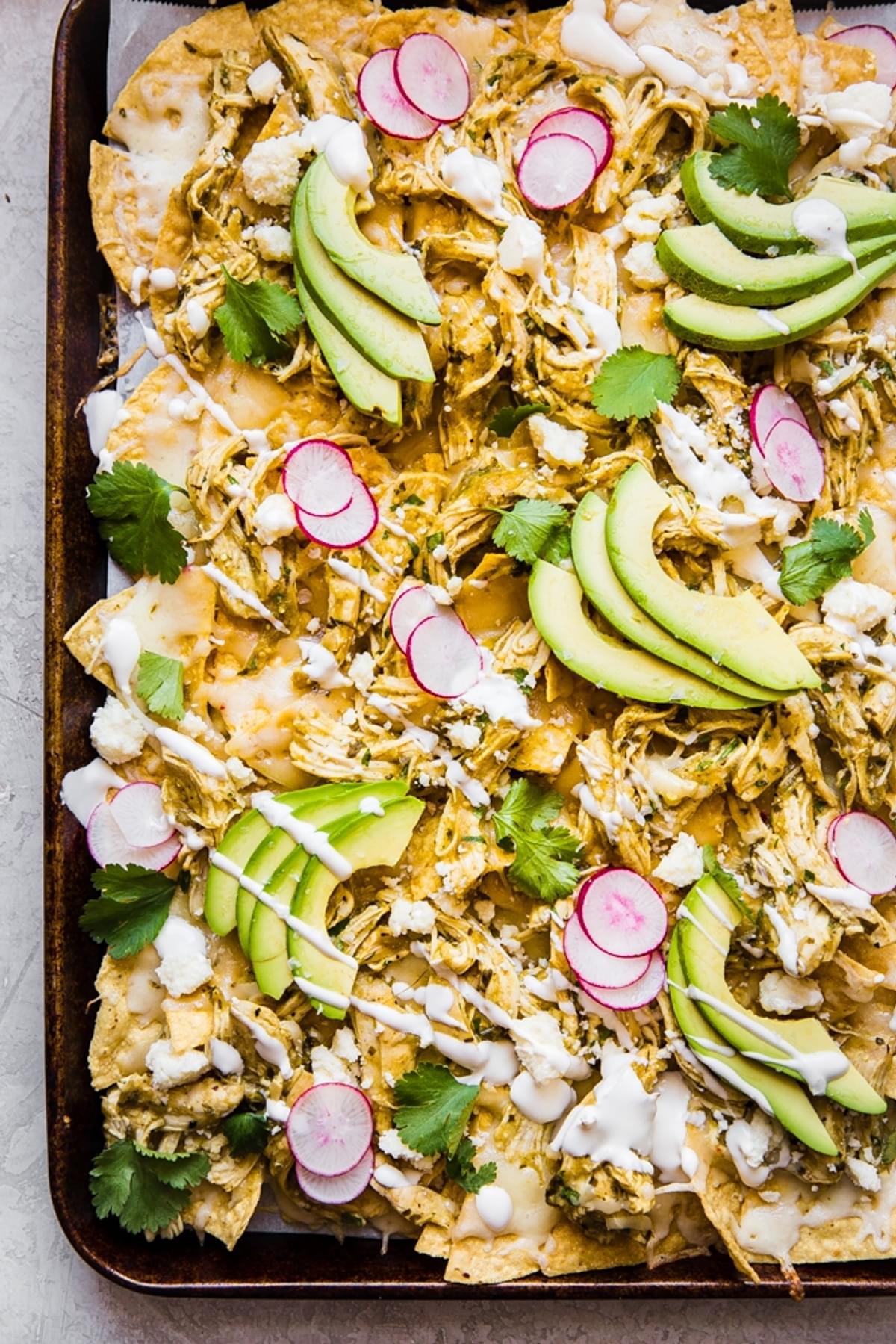 close up of salsa verde nachos shown on a baking sheet topped with avocado, radishes and sour cream