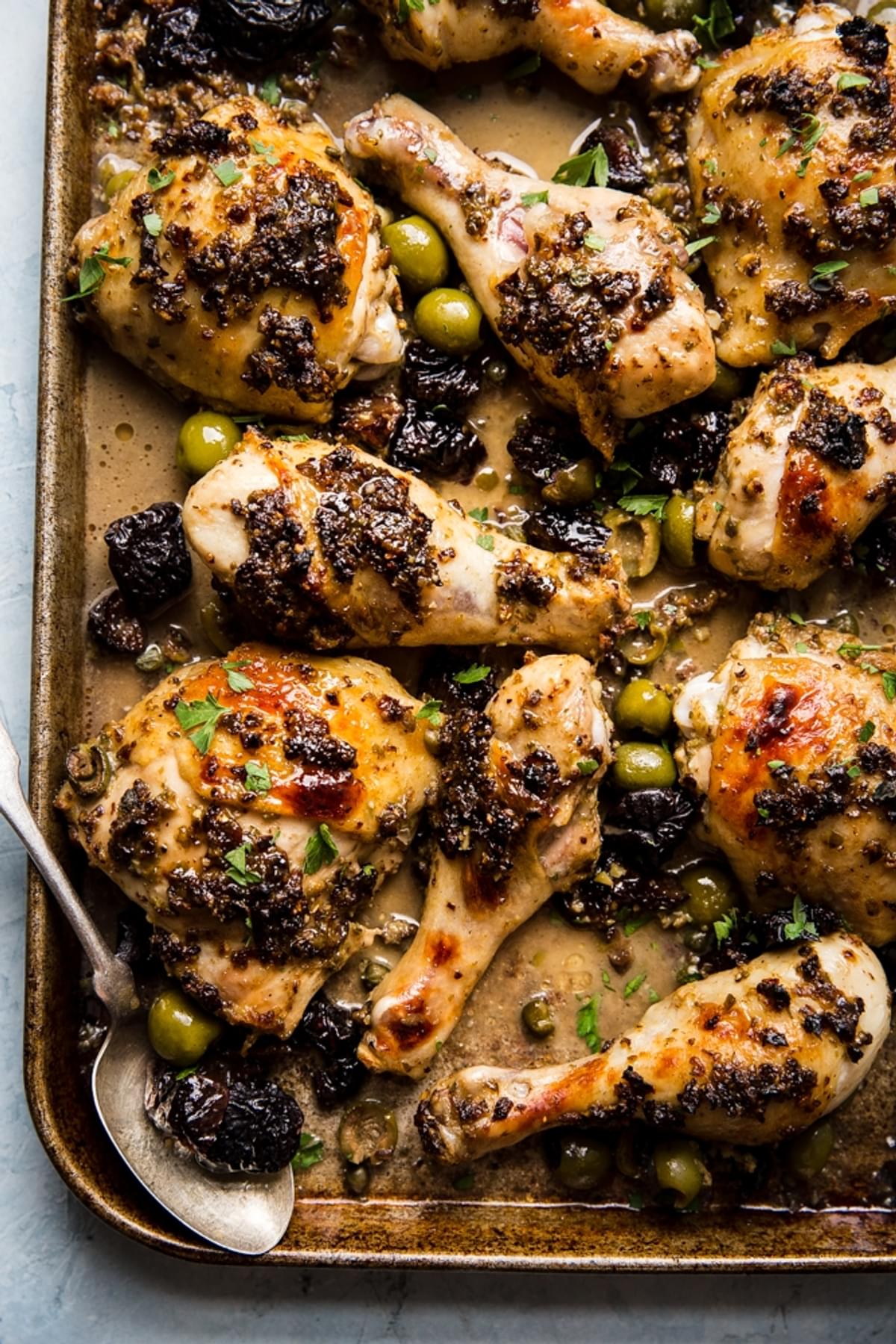homemade chicken marbella with olives, prunes, capers and a pan sauce on a sheet pan with a spoon
