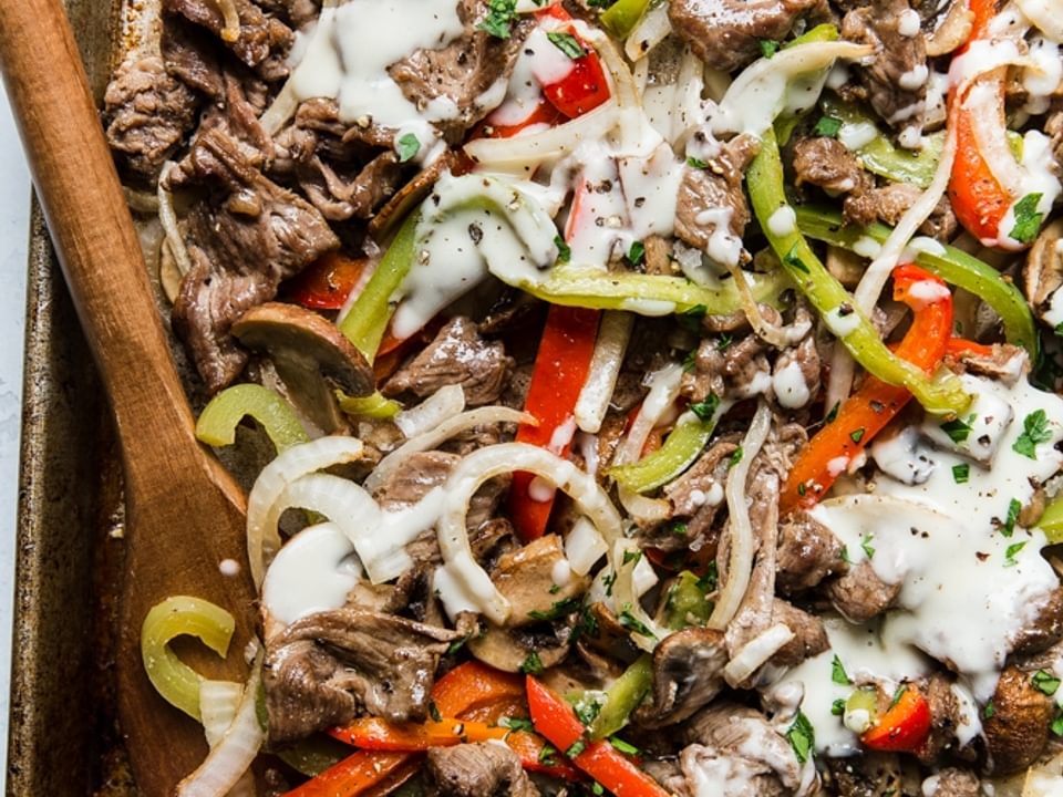 Steak, red and green bell peppers, onions and mushrooms with cheese sauce on a rimmed baking sheet for Philly Cheesesteak