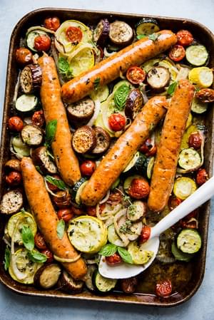 ratatouille baked on a sheet pan with 5 sausage links with a white serving spoon.