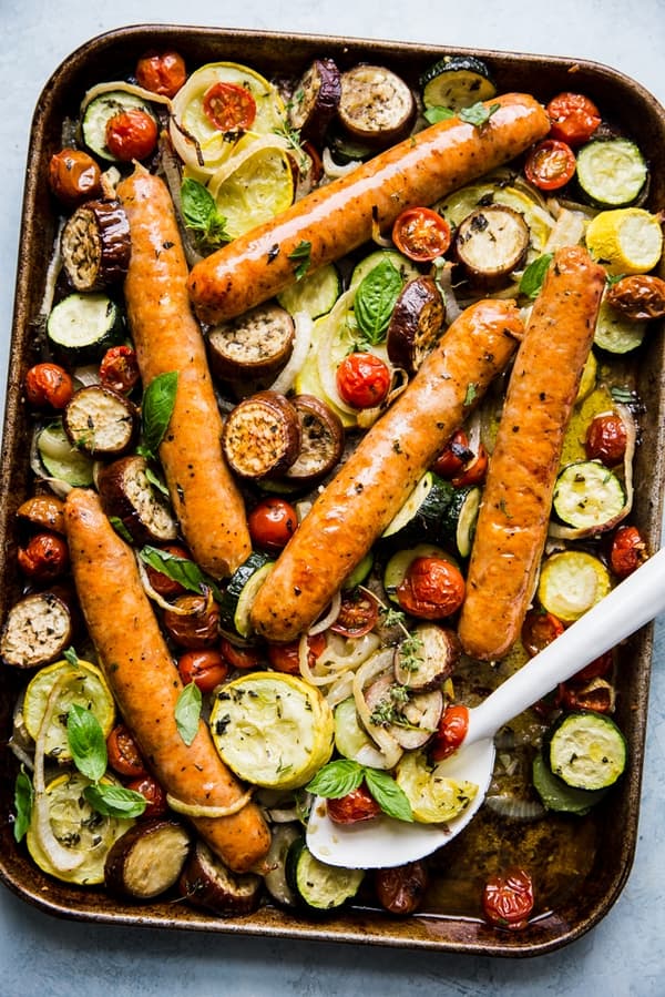 ratatouille baked on a sheet pan with 5 sausage links with a white serving spoon.