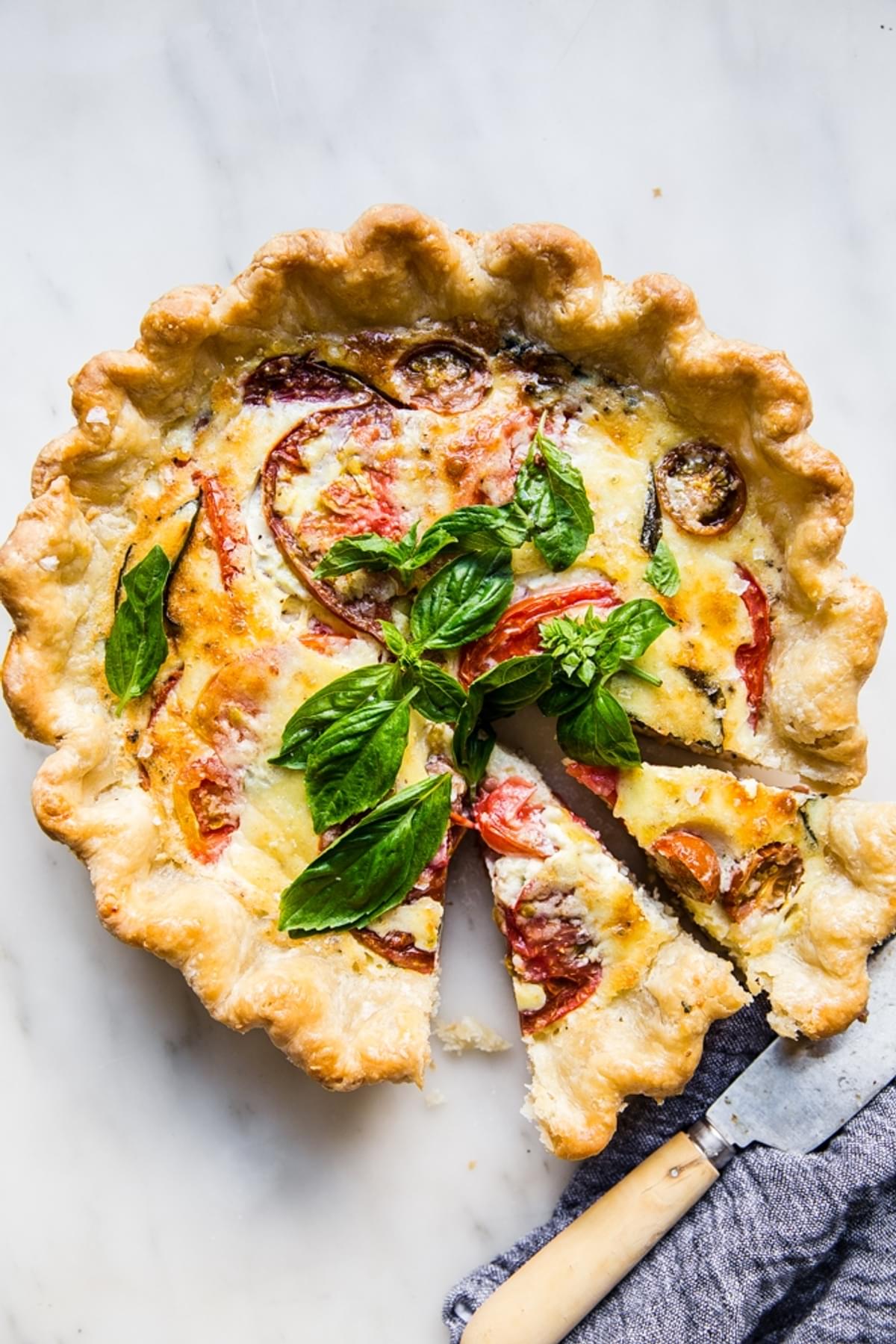 tomato, basil quiche on a marble counter with two slices cut out of it shown next to a knife and linen