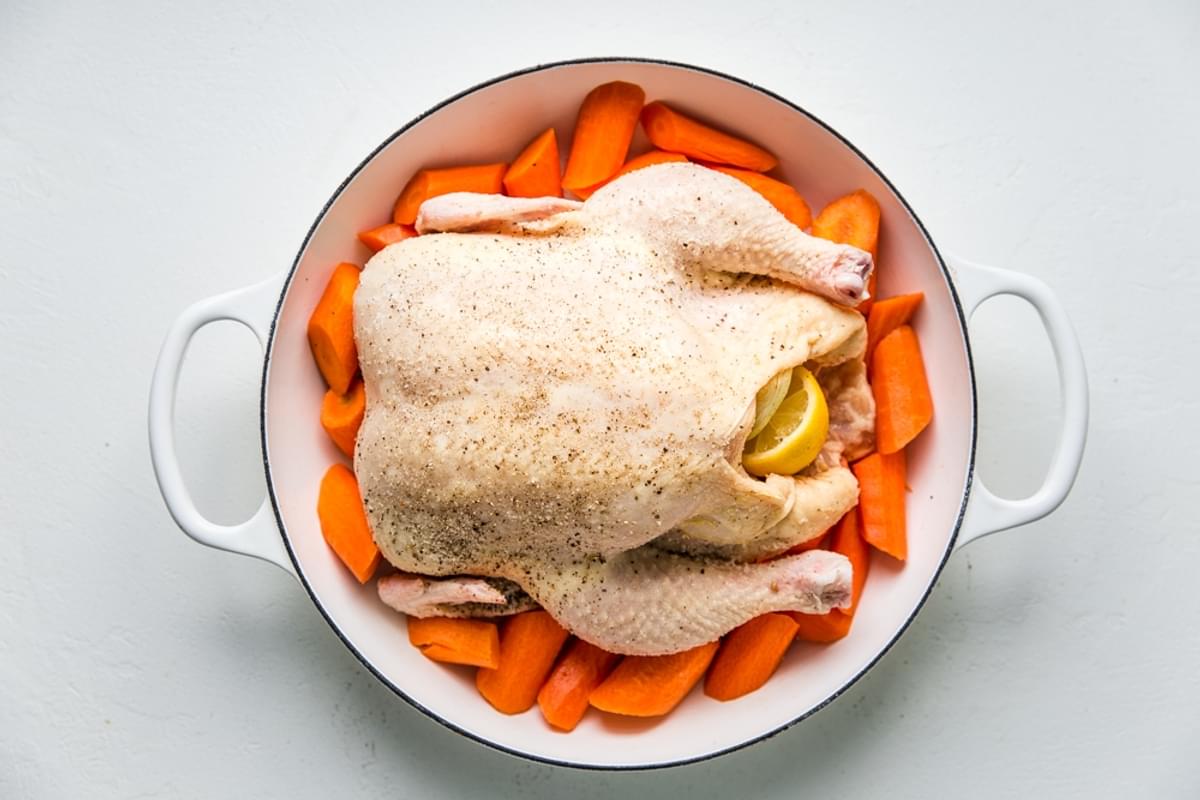 Raw whole chicken shown over carrot slices in a large white braiser