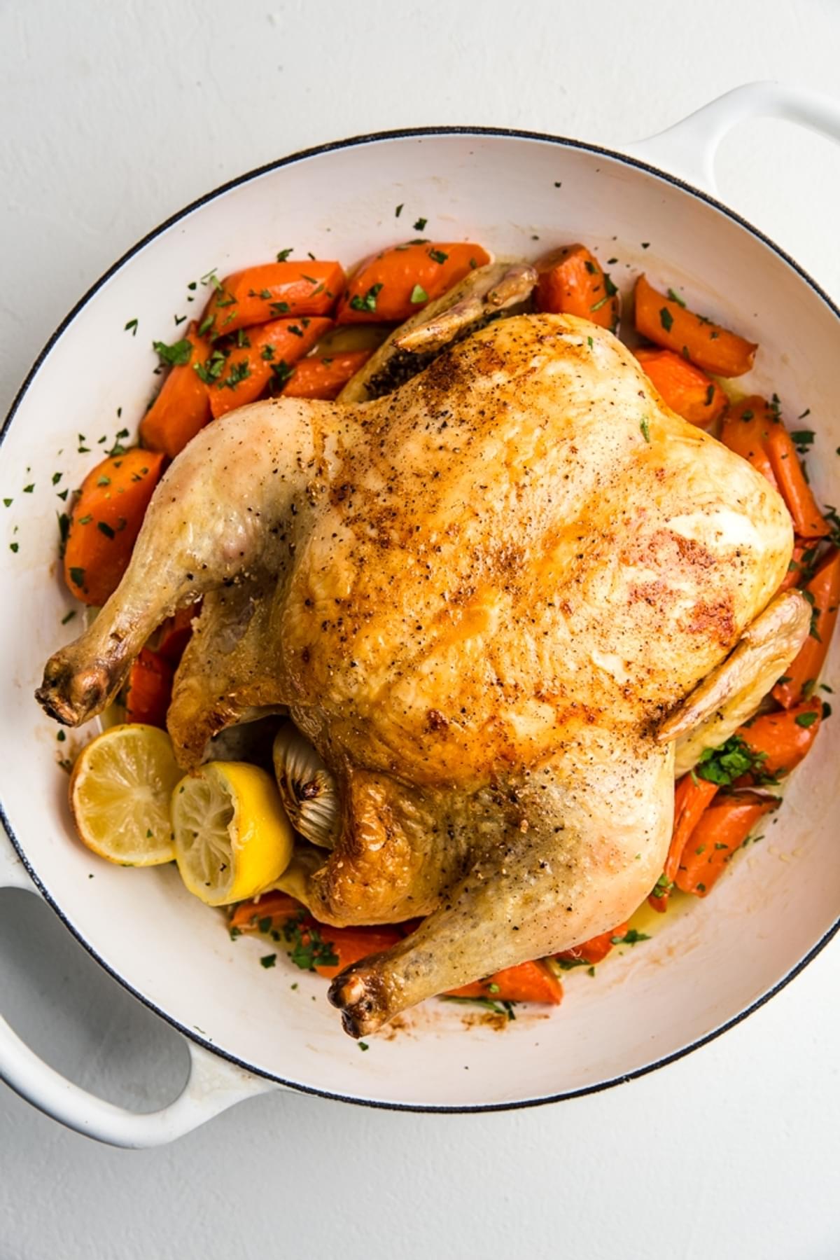 whole roast chicken with carrots shown in a large white braiser with lemon slices