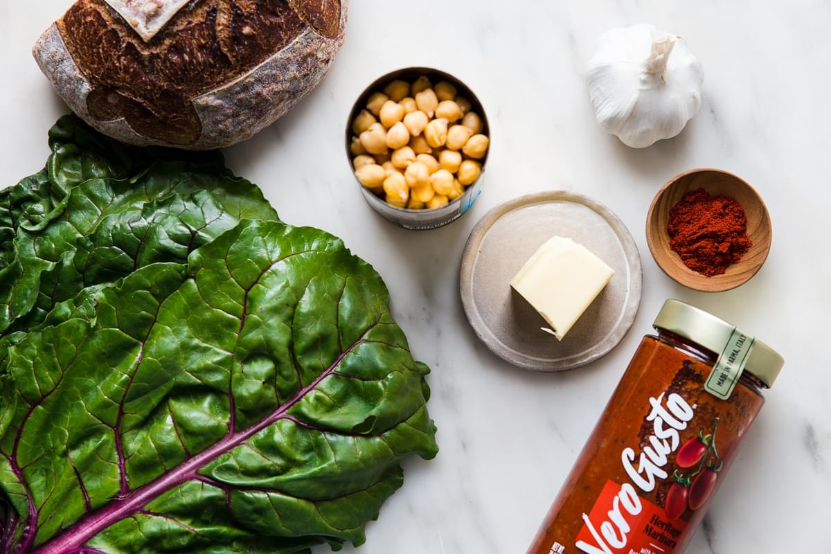 swiss chard, crustly loaf of bread, can of chickpeas, butter, smoked paprika, garlic and a jar of tomato sauce shown.