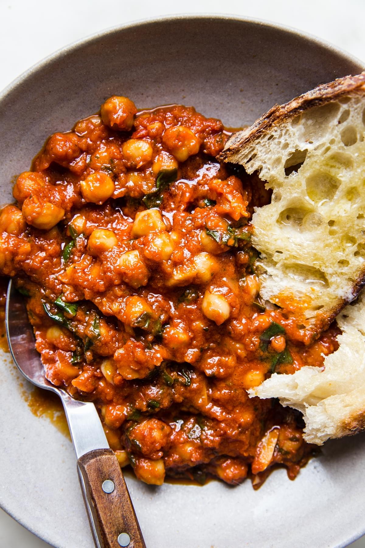 braised chickpeas in tomato sauce with chard shown in a ceramic bowl with sourdough bread and a wooden handled spoon.