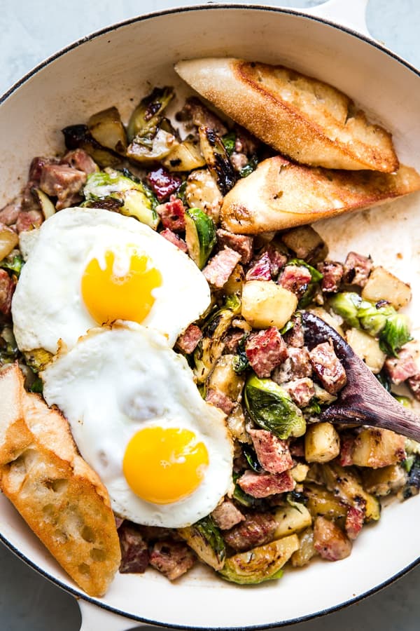 corned beef hash in a skillet topped with 2 fried eggs and served with toasted crusty bread