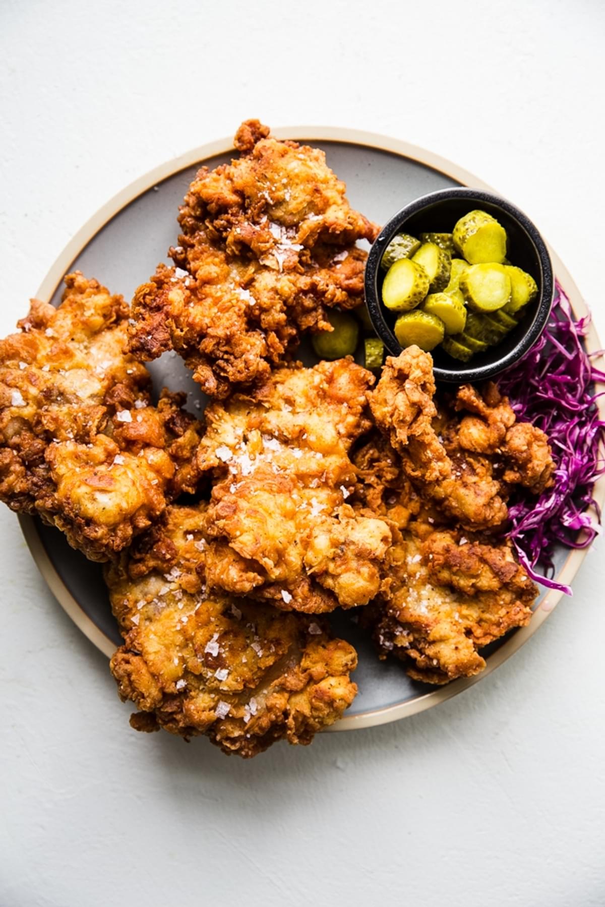 Perfectly crispy fried chicken thighs show on a plate along with a small bowl of pickles and pickled red cabbage.