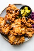 perfectly crispy fried chicken on a platter shown with a bowl of sliced pickles and pickled cabbage.
