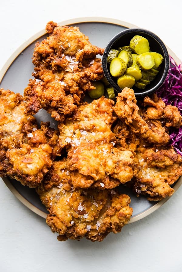 perfectly crispy fried chicken on a platter shown with a bowl of sliced pickles and pickled cabbage.